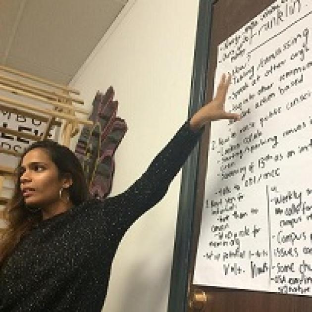 Young black woman with long brown hair and a long sleeved black shirt pointing to a sign up on a wall with lots of black writing on it