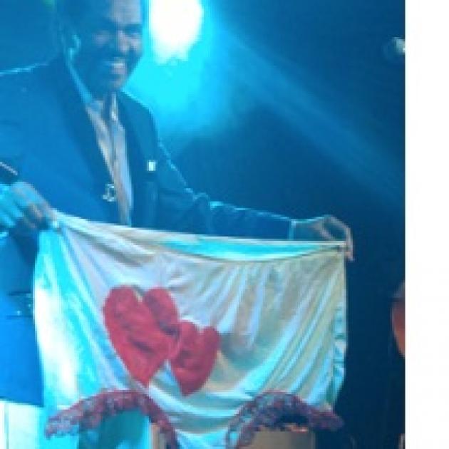 Smiling black man standing in a suit on a stage holding up a huge pair of women's undies that are white with two red harts on them