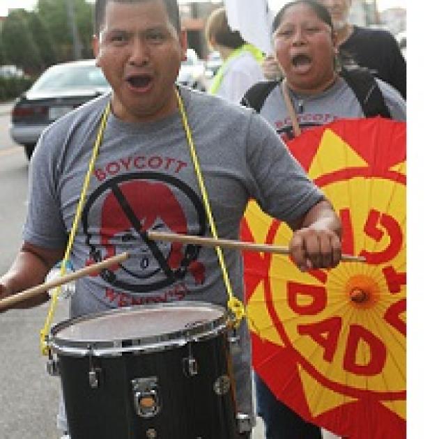 Latino man in foreground with drum around his neck and hitting it with drumsticks, his mouth open chanting, wearing a shirt that says Boycott Wendys with a picture of a girl's head with red pigtails and a No sign around it (circle with line through it) and a Latino woman behind him holding a bright orange and yellow sign also with her mouth open chanting