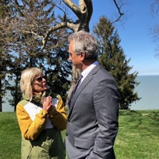 Short blonde wtoman wearing a yellow coat and sunglasses talking and gesturing to a taller white man with gray hair and a gray suit outside by tree and a lake 