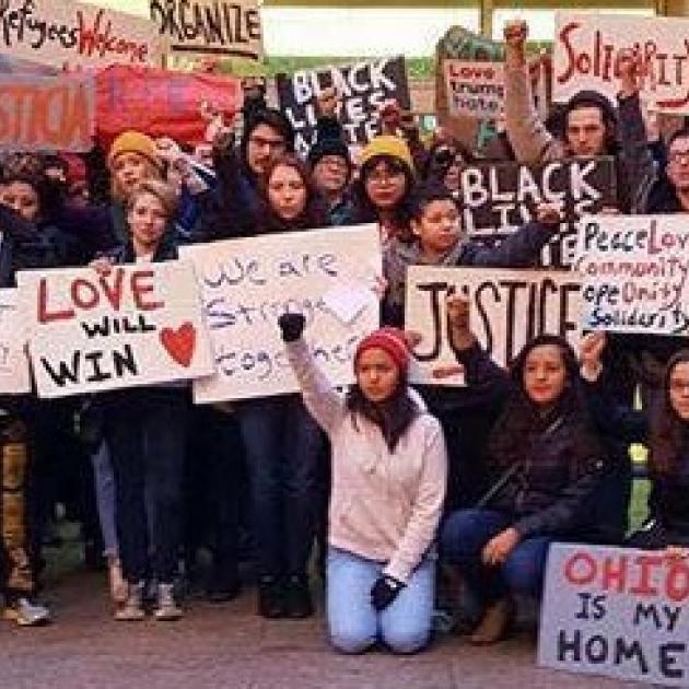 Many people posing in rows outside wearing winter clothes holding signs that say Live Will Win, Refugees Welcome, Justice, Ohio is my Home, Solidarity, Organize, and a woman in front on her knees with a fist in the air