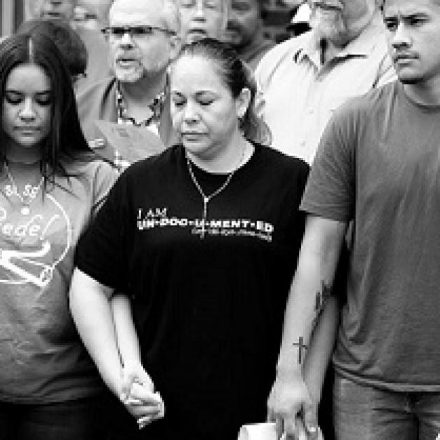 Black and white photo of three Latino people, a young girl with long brown hair looking down, a middle-aged woman with a black T-shirt and necklace looking down and a young man looking very worried, all holding hands with several people behind them 