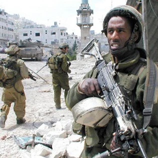 Foreground has a dark skinned man holding as assault rifle in the background two military men with rifles walking toward a building