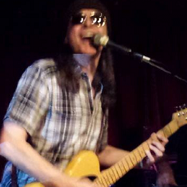 Man with long brown hair in plaid shirt playing a guitar and singing into a mic