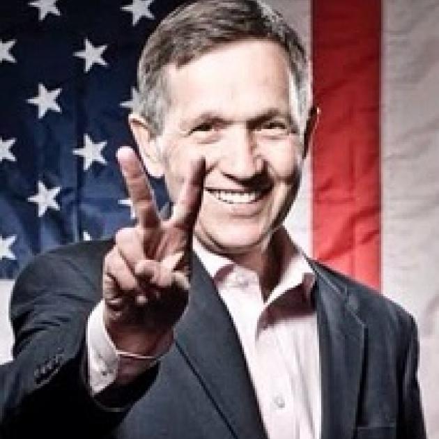 Older man with gray sideburns smiling and holding his fingers in a peace sign with a flag in the background