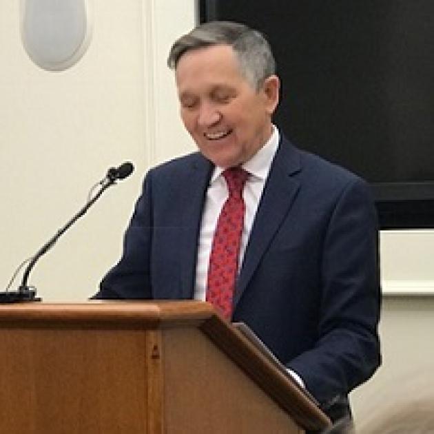 Older white man in suit with red tie at a brown podium looking down, smiling and talking into a mic