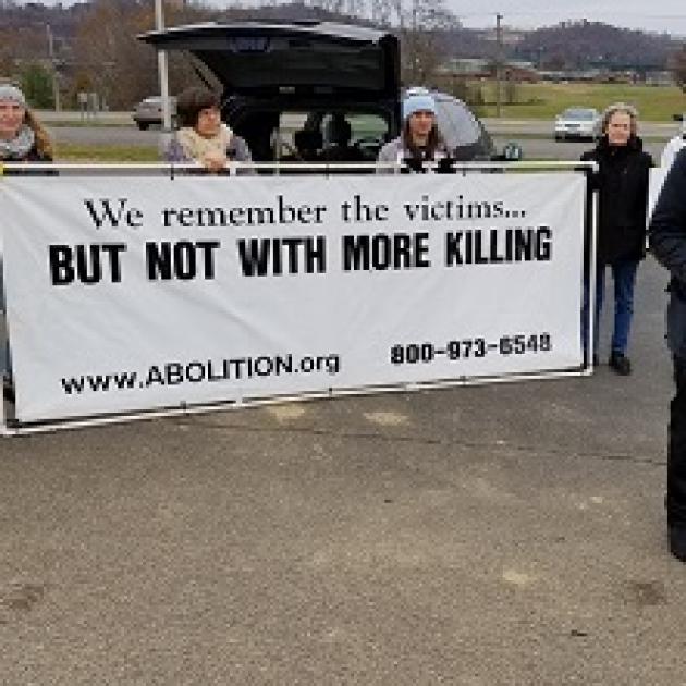 People outside holding a long white banner with black letters saying We remember the victims...But not with more killing and www.abolition.org and a phone number