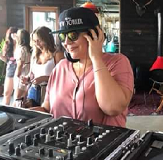 Woman with a baseball cap on and headphones at a mixing board smiling, with people in the background