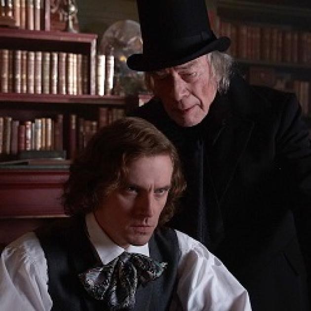A man with chin length curly brown hair looking intense sitting down with old fashioned clothes from the 30s and an older gray haired man in black with a black top hat leaning over his shoulder saying something in his ear, bookshelves in the background