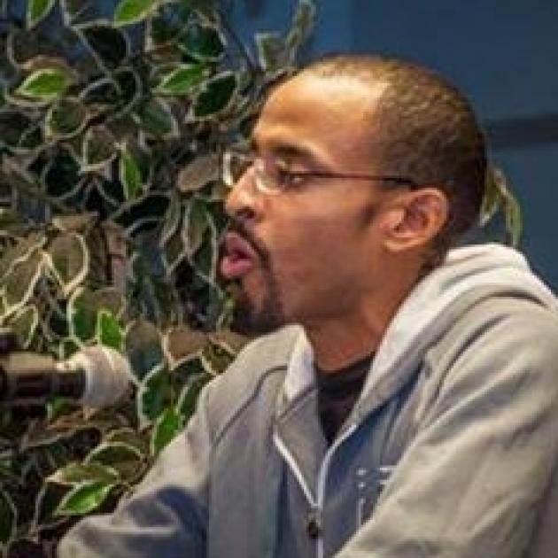 Young black man facing left talking into a microphone, he has short cropped hair and glasses and a goatee, sitting in front of a plant