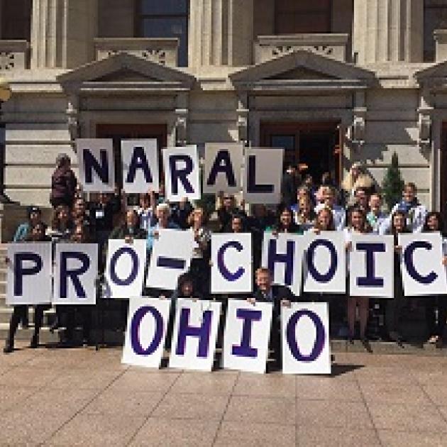 Big white capitol building with columns and turrets above windows in the background, lots of people posing in front holding placards each with a letter spelling out NARAL PRO-CHOICE OHIO