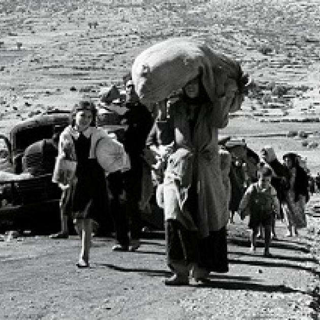 Black and white photo of women and children walking down a dusty path outside with the woman in the foreground carrying a big heavy bag on her head