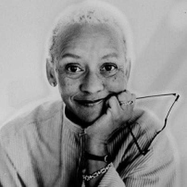 Black and white photo of older black woman head and shoulders smiling with her glasses in her hand with her chin in her hand. 