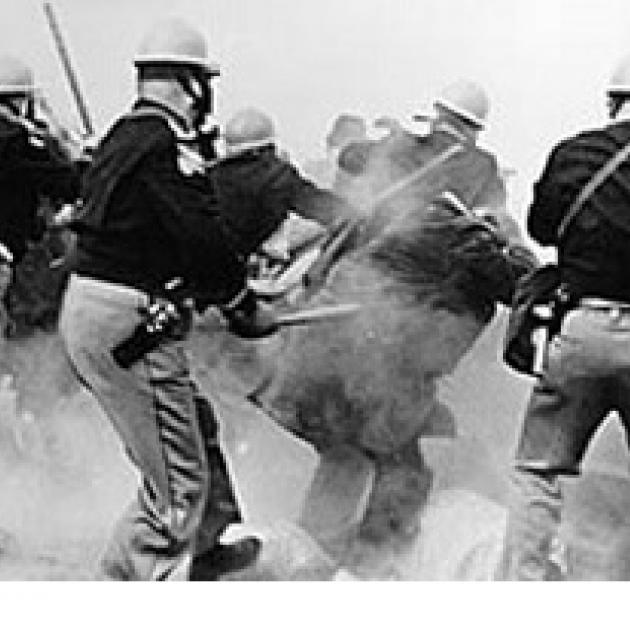 Black and white photo of police wearing helmets with their backs to the camera, looking like the are beating people with sticks among a lot of gas in the air obscuring the people in the picture