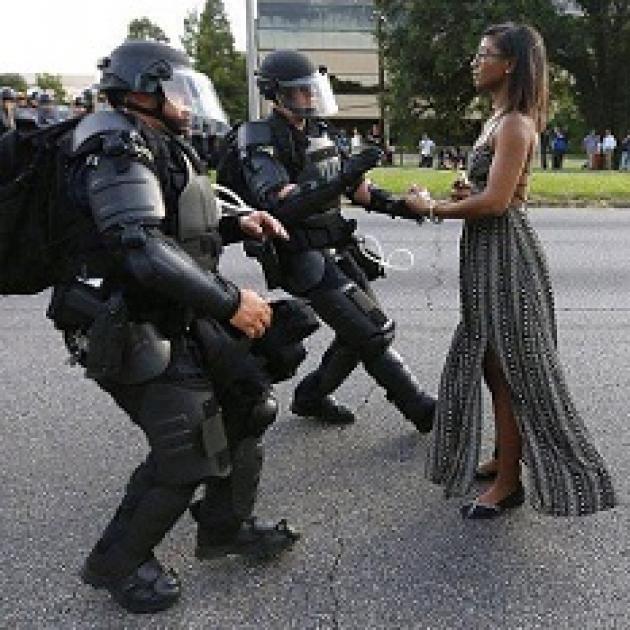 Young black woman in long flowing black and white dress wearing glasses standing straight and tall and calm and two older white heavily armed riot police looking like they are stepping back away from her