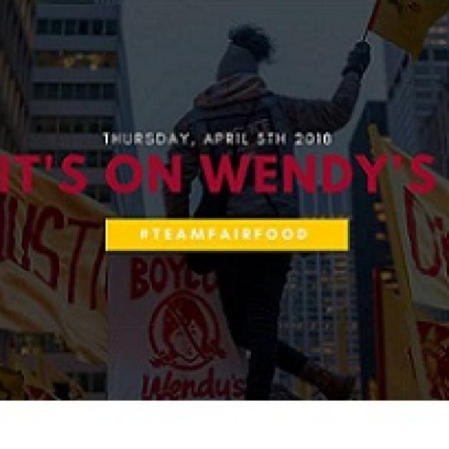 Darkened photo of back of a black woman outside waving a yellow flag over her head and signs all around her about justice and against Wendys