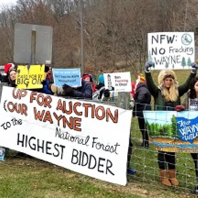 Lots of young people wearing coats outside near a forest at a fence with protest signs reading Up for Auction Our Wayne to the Highest Bidder