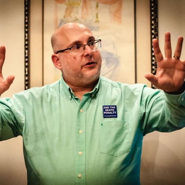 White middle aged guy with glasses holding his arms in the air with fingers spread on hands while he talks and wears an Abolish the Death Penalty button