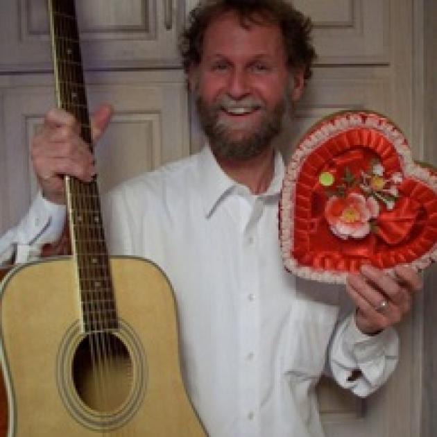 Man with a beard and a big smile and wearing a white button down shirt standing in front of a door holding a guitar and a red heart shaped box