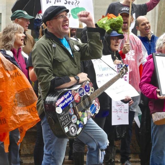 Guy with cap and guitar covered with stickers with his fist in the air in front of a crowd of people