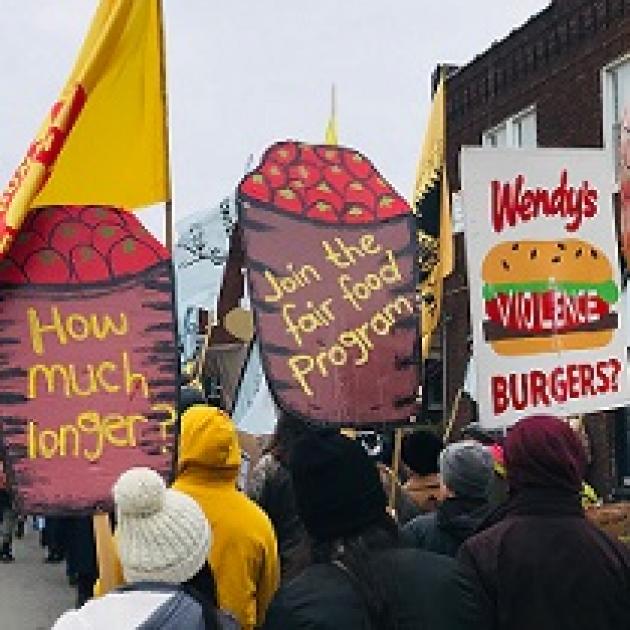 People marching with very large high signs with depictions of barrels of tomatoes with the words How much longer? and Join the Fair Food program and a Wendy's hamburger that says violence on it