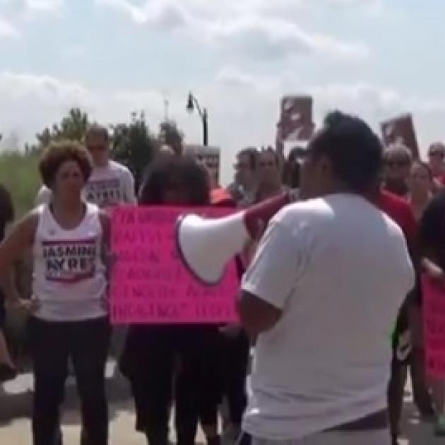 Man with brown hair and white shirt with back to camera holding a bullhorn surrounded by protestors