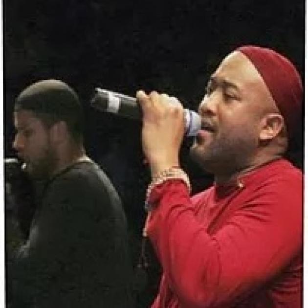 Black men in concert, one in red singing into mic