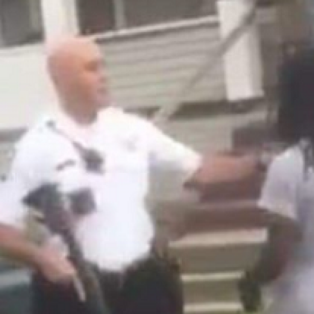 Blurry photo of a white bald policeman in a white shirt and holding a rifle pushing a black man with long braids