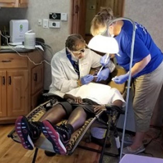 Person in a dental chair and a dentist to the left working on them and a woman to the right, both with mask and gloves on