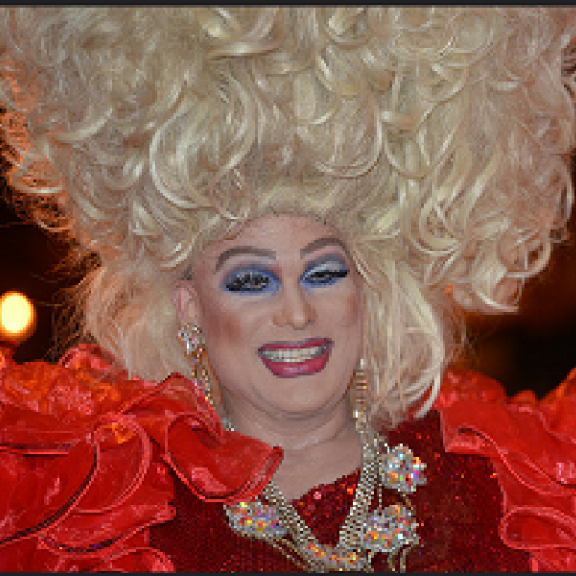 Man dressed a woman with huge blonde hair in curls going up in the air lots of blue eye shadow, red lips and a red outfit