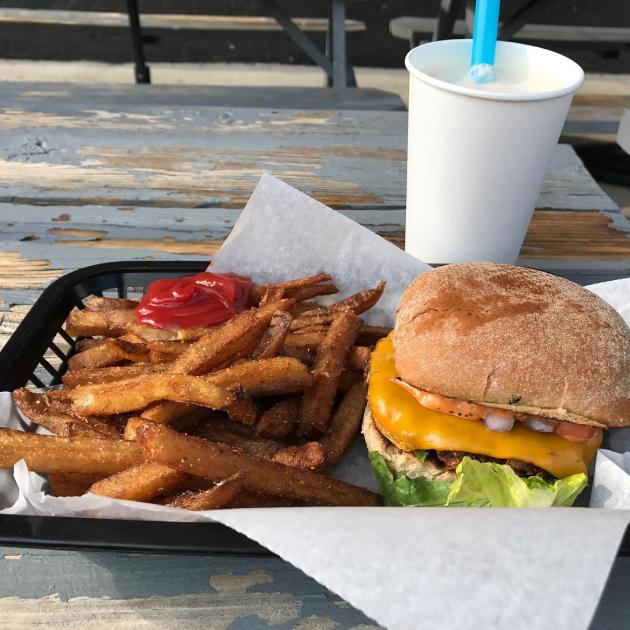 A tray on a wooden table outside with a white cup and blue straw, a bunch of french fries and a burger with visible lettuce and cheese