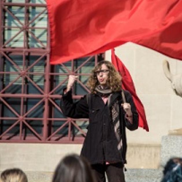Young white man with long brown curly hair and glasses holding a very large red flag standing in front of a crowd raising his fist in the air