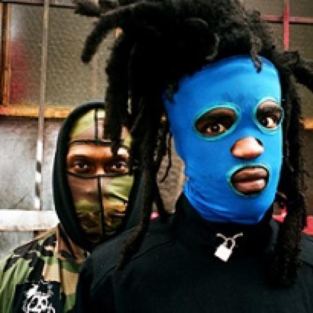 Two men one in front with dreds sticking up off his head and a bright blue mask and a guy behind him with a camouflage mask