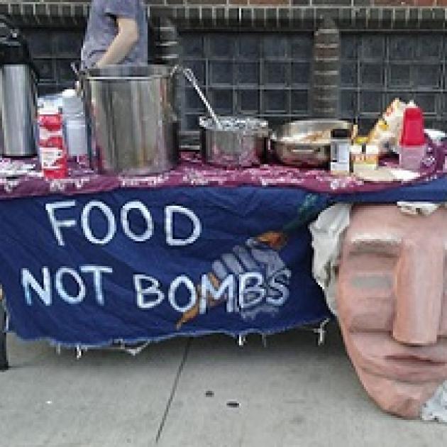 Big paper mache face hanging on the front of a table outside next to a blue sign with white hand drawn letters that say Food Not Bombs and a lot of pots and pans and condiments on the table and a brick wall in the background