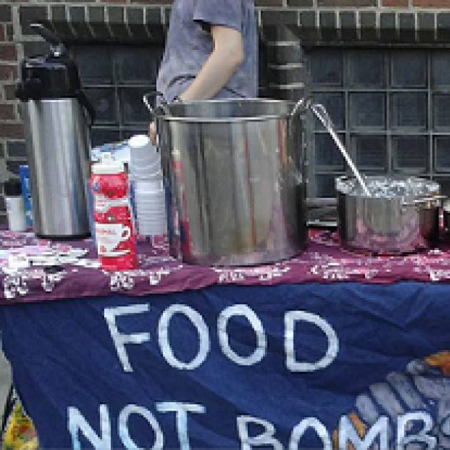 Outside table with pots and pans on it and tablecloth with sign Food not Bombs
