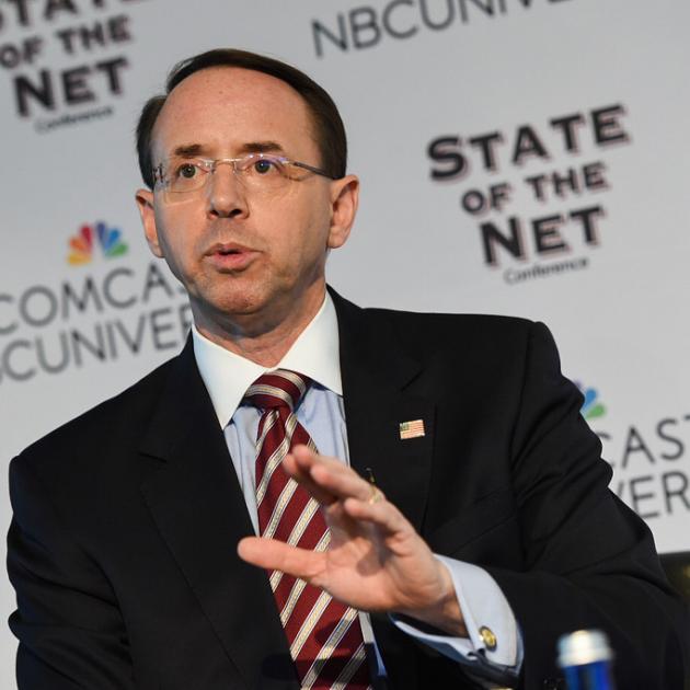 Balding white man with wire rimmed glasses wearing a suit speaking and make a hand gesture
