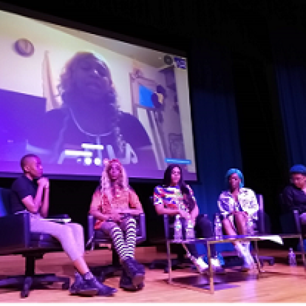 Stage with large screen with a black trans woman speaking and five black women in chairs below