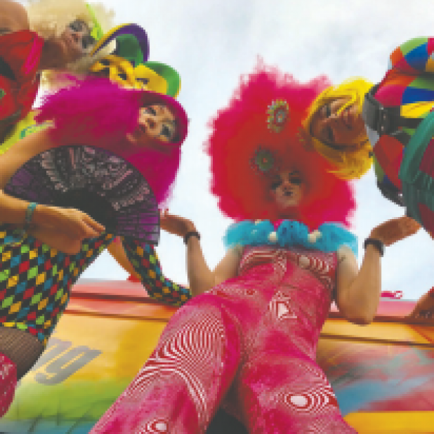 Camera angle looking from ground up at four decked out women with bright orange, pink and rainbow colored fluffy feathery bedazzled outfits