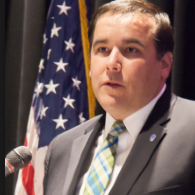 Middle aged white man in a suit with brown receding hair at a mic with a flag in the background