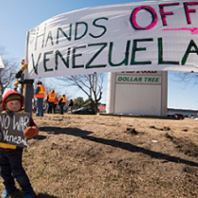 Big banner outside saying Hands off Venezuela and very small boy holding sign saying No War Venezuela