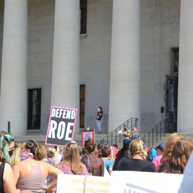 crowd protesting in front of state house with sign that says defend roe