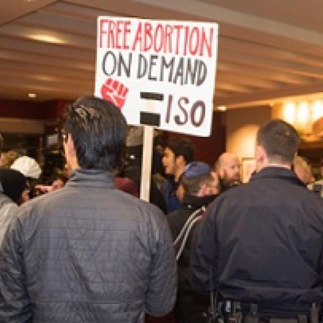 Backs of two men, one with a cop looking uniform on and the other with a sign that says Free Abortion on Deand ISO with a red fist