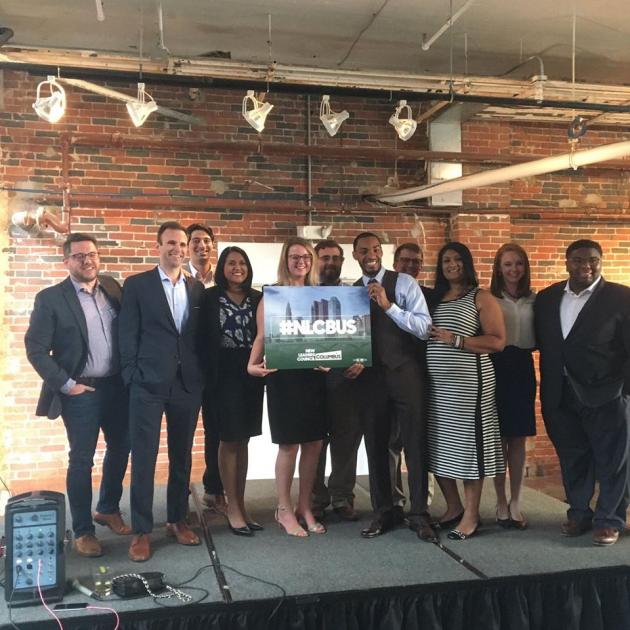 A big room with lights hanging from the ceiling and a group of white and different people of color smiling and holding a poster of the Columbus skyline