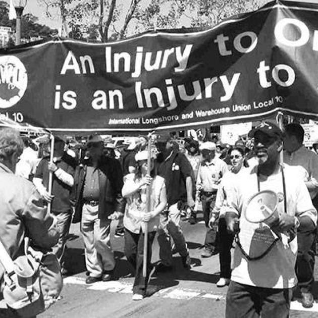 Black and white photo of big banner with words An Injury to one is an injury to all and mostly black people marching with it, some with drums