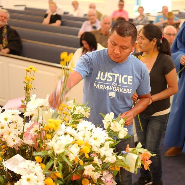 Main in a Justice for Farmworkers light blue T-shirt putting flowers into a huge container that has a lot of flowers in it and a line of people behind him and pews of a church behind them