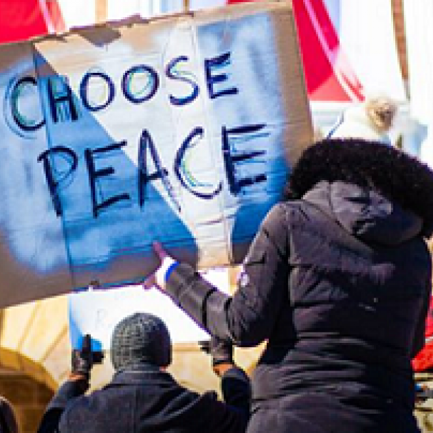 Woman with her back to the camera wearing a large winter coat with furry hood holding a big sign that reads CHOOSE PEACE, she is with a group of people at a rally