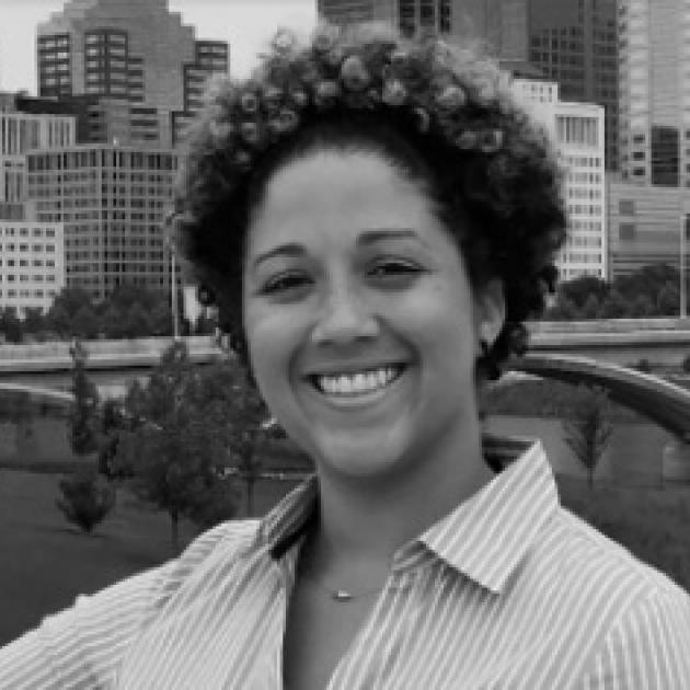 Smiling black woman with short cropped curly hair and a striped button down shirt standing with downtown buildings and a bridge across the river behind her