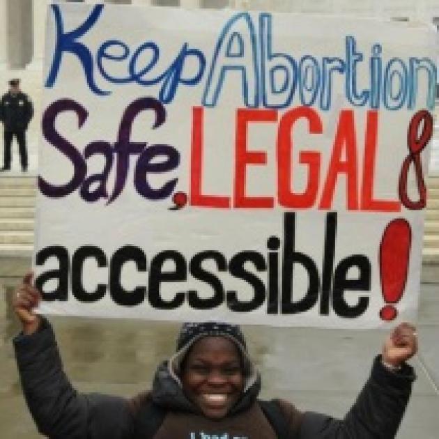 Black woman holding sign saying Keep abortion safe, legal and accessible in from of a government building with a cop standing in the background