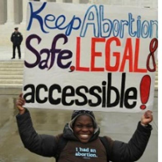 Young black woman smiling with winter clothes on outside an official looking government building holding a sign above her head saying Keep Abortion Safe, Legal & accessible! A cop is standing back in the distance.