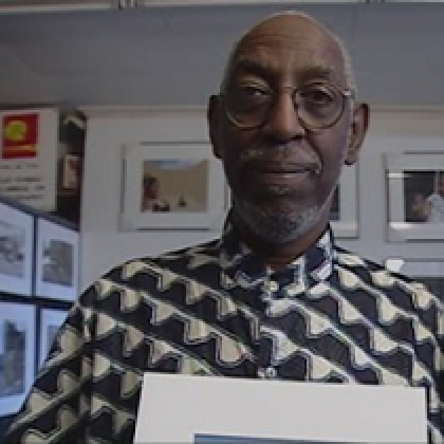Older black man, bald with gray mustache and beard in a gray and white shirt holding a photo and standing in front of lots of other photos on the wall. 
